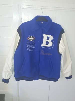 Blå och vit collegejacka  - Säljer en blå och vit bomberjacka ifrån H&M. Stängs med knappar framtill och har två fickor. Perfekt för en sportig och trendig look.