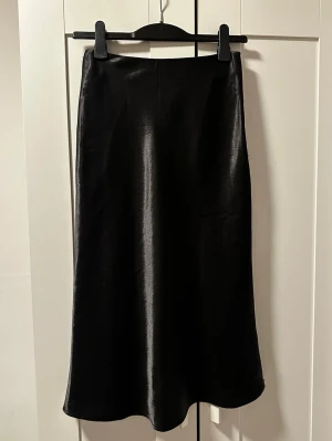 Svart långkjol i satin-look - Säljer en svart långkjol från H&M med glansig satin-finish. Kjolen har en enkel, rak siluett och slutar vid ankeln. Perfekt för dig som gillar stilrena och trendiga plagg. Kjolen har en diskret dragkedja i sidan för enkel på- och avtagning.