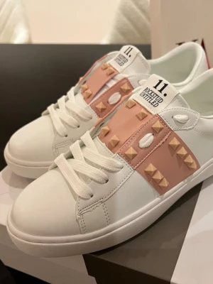 Valentino Rockstud sneakers i vitt och rosa - Snygga Valentino Rockstud sneakers i vitt skinn med ljusrosa paneler och ikoniska nitar. Skorna har rund tå, platt sula och snörning framtill. Märkeslogga på plös och sula, samt detaljerade nitar runt hälen och på sidorna. Perfekt för dig som gillar statement-detaljer.  Finns i stl. 38,39