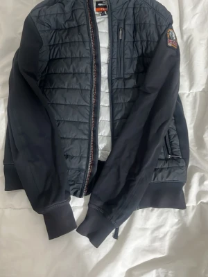 Svart quiltad bomberjacka Parajumpers - Säljer en svart quiltad bomberjacka från Parajumpers i storlek Y-M. Jackan har ribbade muddar, dragkedja framtill, bröstficka med dragkedja och patch på ärmen. Materialet är polyester och fleece, vilket gör den både lätt och varm.