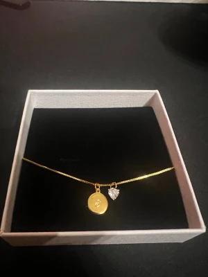 18K guldpläterat halsband med berlock - Snyggt och stilrent halsband i 18K guldplätering med en rund bricka graverad med bokstaven E och ett gnistrande hjärthänge i kristall-look. Kedjan är tunn och elegant, perfekt för att lyfta vilken outfit som helst. Levereras i en vit presentask.   Helt oanvänd. Nypris 1198kr