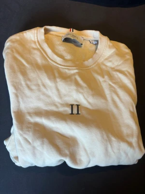 Vit sweatshirt från Les Deux - Säljer en stilren vit sweatshirt från Les Deux med svart 'II'-brodyr på bröstet. Tröjan har rund halsringning, ribbade muddar och är tillverkad i mjuk bomull. Perfekt för dig som gillar minimalistisk design och vill ha något bekvämt till vardags.