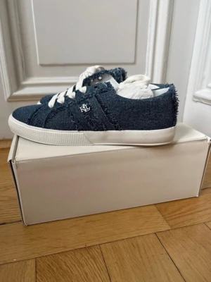 Blå denim sneakers från Lauren Ralph Lauren - Säljer ett par snygga sneakers från Lauren Ralph Lauren i blå denim med råa kanter och vita snören. Skorna har vit sula och RL-logga på sidan. Perfekta för dig som gillar en avslappnad men trendig stil. Skorna är köpta för 1500kr och alldrig andvända!!