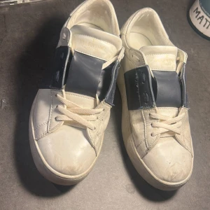 Valentino Garavani sneakers vit/blå - Snygga sneakers från Valentino Garavani i vitt skinn med bred mörkblå rem över snörningen och coola nitar på hälen. Klassisk rund tå och platt sula, perfekt för dig som gillar exklusiv streetstil. Skorna har vita snören och en chunky siluett.