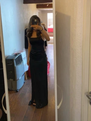 Svart långklänning med smala axelband - Säljer en elegant svart långklänning med smala axelband och rak urringning. Klänningen har en figurnära passform och är tillverkad i ett glansigt material som ger en lyxig känsla. Perfekt för speciella tillfällen när du vill känna dig extra snygg.