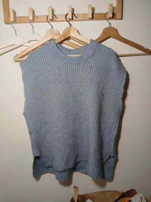 Blå stickad ärmlös tröja - Säljer en blå, grovstickad ärmlös tröja med rund hals. Perfekt att bära över en skjorta eller t-shirt för en chill lager-på-lager-look. Tröjan har en avslappnad passform och ribbade kanter vid hals och nederkant.