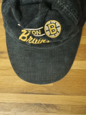 Grå Boston Bruins manchesterkeps - Säljer en grå keps i manchester med broderad Boston Bruins-logga och text framtill. Kepsen är officiellt licensierad av NHL och har en klassisk böjd skärm. Perfekt för dig som vill representera ditt favoritlag med stil.