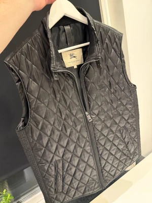 Svart väst från Burberry - Säljer en svart väst från Burberry med dragkedja framtill och ståkrage. Västen har två fickor och klassiskt rutmönster i quiltad design. Perfekt för lager på lager och snygg till både hoodie och skjorta. Ända grejen är att knappen saknas (se bild) annars i väldigt bra skick, pris kan diskuteras