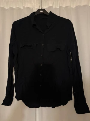 Svart skjorta från 157 med fickor - Svart skjorta från 157 i storlek L. Skjortan har två bröstfickor med lock, klassisk krage och knäppning framtill. Långärmad modell i mjukt tyg som passar till många olika stilar.