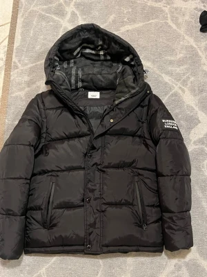Svart pufferjacka från Burberry - Säljer en svart pufferjacka från Burberry med avtagbara ärmar så den kan användas som väst. Jackan har huva med rutigt foder, dragkedja och knappar framtill samt två fickor med dragkedja. Perfekt för kalla dagar och riktigt snygg streetstil. Helt ny utan prislapp. Det är en 🐄Pia.