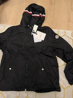 Svart vindjacka från Moncler - Säljer en svart vindjacka från Moncler med huva och dragkedja framtill. Jackan har vita och röda ränder på huvan, två fickor med tryckknappar och Moncler-logga på ärmen. Tillverkad i lätt polyester, perfekt för blåsiga dagar.