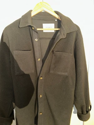 Grå fleece overshirt från Les Deux - Säljer en grå fleece overshirt från Les Deux med två bröstfickor och knappar framtill. Jackan har en mjuk, teddy-liknande yta och klassisk krage. Perfekt lager på lager-plagg för kyliga dagar. Broderad Les Deux-logga på ena fickan och diskret detalj i nacken.