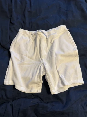 Vita shorts från BOSS Homewear - Säljer ett par vita shorts från BOSS Homewear i mjukt bomullstyg med ribbad struktur. De har snörning i midjan och en relaxed passform. Perfekta för chill dagar eller när du vill ha det bekvämt hemma.