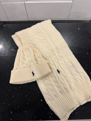 Ralph lauren mössa halsduk - Bilderna visar en stickad mössa och halsduk från Polo Ralph Lauren i cremevitt, med klassiskt kabelstickat mönster och blå logga. Ingen väst syns på bilderna.
