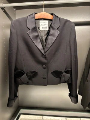 Svart kavaj med satindetaljer Moschino - Säljer en svart kavaj från Moschino med satinkrage och dekorativa rosetter i satin vid fickorna. Kavajen är croppad, har klädda knappar framtill och långa ärmar med satinkant. Perfekt för dig som vill ha en unik och elegant look.