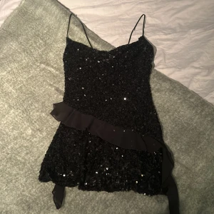 Svart paljettklänning från Zara - Supersnygg svart kort klänning från Zara med tunna axelband och glittriga paljetter över hela tyget. Klänningen har en volangdetalj diagonalt över framsidan som ger extra edge. Perfekt för dig som vill sticka ut och glänsa.