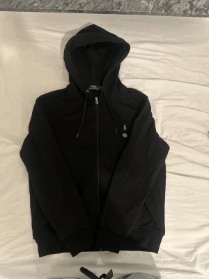 Ralph lauren zipper hoodie - Svart zipper hoodie i storlek M, i mycket bra skick. Säljs eftersom jag har vuxit ur den. Fick den i present förra året och den är sparsamt använd. Tillverkad i 69 % bomull och 31 % polyester, vilket gör den både bekväm och slitstark. En klassisk och stilren hoodie som är väldigt populär. Nypris från Zalando är cirka 2135 kr. Är öppen för prisförslag!😊