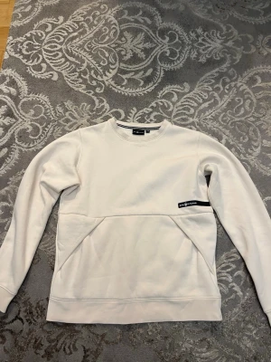 Vit tjocktröja från Sail Racing - Säljer en stilren beige tjocktröja från Sail Racing med svart logotyp på bröstet och ett tryck i nacken. Tröjan har lång ärm, rund hals och praktisk ficka framtill. Materialet är mjuk bomull och polyester, perfekt för kalla dagar!
