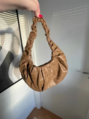 Brun axelväska  - Trendig brun axelväska från Nelly. Väskan har en scrunchie-inspirerad axelrem och en rundad, puffig form. Oanvänd 