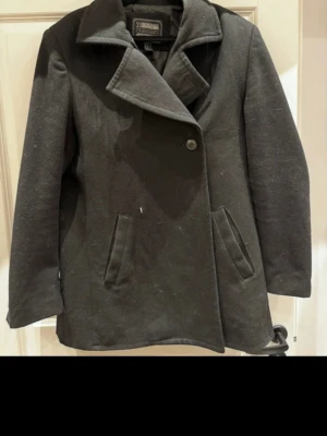 Svart ullkappa från 21MEN svartfärgad storlek M - Stilren svart kappa från 21MEN i ullblandning. Klassisk design med bred krage, två snedställda fickor och knäppning framtill. Fodrad insida och fyra knappar vid ärmslutet ger en snygg touch. Perfekt för kyligare dagar.