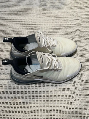 Nike Air Max 270 vita sneakers - Säljer ett par Nike Air Max 270 sneakers i vitt med svarta detaljer. Skorna har en stor synlig Air-enhet i hälen, mesh-ovandel och snörning. Snygg och sportig design med svart sula och logga på sidan. Perfekta för dig som gillar streetwear och bekväma skor.