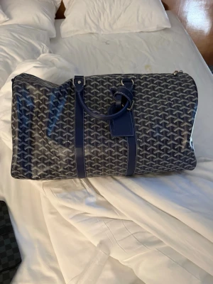 Goyard väska  - Säljer en stor och rymlig blå resväska från Goyard med deras ikoniska grafiska mönster. Väskan har två handtag, detaljer i blått skinn och dragkedja upptill. Perfekt för weekendresor och har en riktigt snygg och unik look.