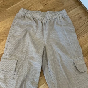 Beige manchester cargobyxor med resårmidja - Säljer ett par beige cargobyxor i manchester med resår i midjan och stora fickor på sidorna. Perfekta för en avslappnad och trendig stil. Byxorna har en loose passform och är riktigt sköna att bära.