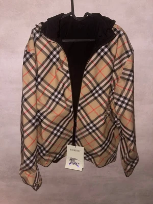 Rutig Burberry vindjacka med huva - Säljer en snygg vindjacka från Burberry med klassiskt rutig mönster i beige, svart, vit och röd. Jackan har huva, dragkedja framtill och Burberry-logga broderad på bröstet. Perfekt för dig som vill sticka ut med stilren design och ikoniskt mönster.