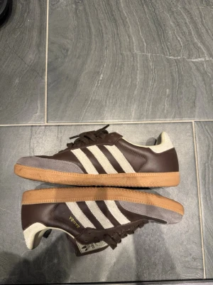 Adidas Samba  - Säljer ett par klassiska Adidas Samba sneakers i brunt skinn med vita detaljer och beige mocka på tån. Skorna har de ikoniska tre ränderna på sidan, gummisula och snörning. Perfekta för dig som gillar retrostil och streetwear.