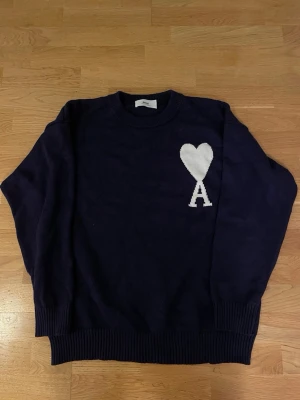 Mörkblå stickad tröja från Ami - Snygg mörkblå stickad tröja från Ami Alexandre Mattiussi med vit hjärt- och A-logga på bröstet. Tröjan har rund hals, ribbade muddar och lång ärm. Perfekt för dig som gillar stilrena detaljer och vill ha något som sticker ut lite.
