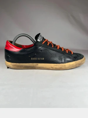 Golden Goose Superstar sneakers svart/röd - Äkta Golden Goose Superstar sneakers i svart skinn med röda detaljer på hälen och vita snören. Storlek 41, orginal snörerna är de röd gula men har även ett par vita om man tycker det är snyggare) som syns på sista bilden.
