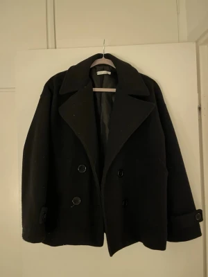 Svart skepparkavaj från Design by Si - Stilren svart skepparkavaj från Design by Si med bred krage och dubbelknäppning framtill. Jackan har stora knappar, klassisk passform och är fodrad på insidan. Perfekt för dig som vill ha en tidlös och snygg look.