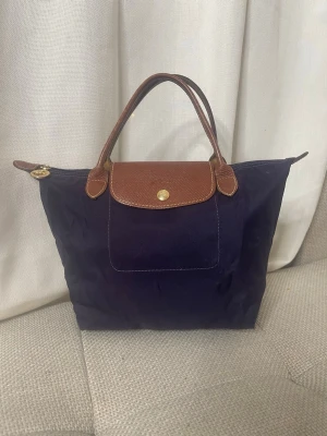 Mörkblå handväska från Longchamp - Säljer en klassisk mörkblå handväska från Longchamp med bruna skinnhandtag och lock. Väskan har guldfärgade detaljer och dragkedja med Longchamp-logga. Perfekt storlek för vardagens essentials och enkel att matcha till olika outfits.
