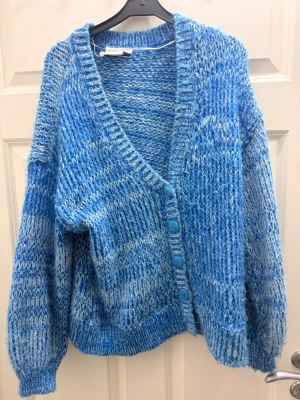 Blå stickad kofta från Vila - Mysig blåmelerad stickad kofta från Vila med v-ringning och stora matchande knappar framtill. Koftan har en loose passform och långa ärmar, perfekt för lager på lager under kyliga dagar.