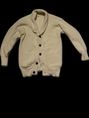 Beige stickad kofta med stora knappar - Säljer en beige stickad kofta med grov struktur och stora svarta knappar framtill. Koftan har två fickor, bred krage och långa ärmar. Perfekt för lager på lager under kyliga dagar.