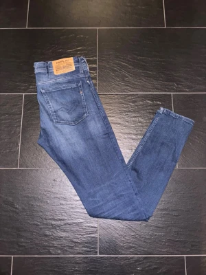 Replay Jondrill blå jeans, storlek 31 - Säljer ett par Replay Jondrill jeans i klassisk blå tvätt. De har fem fickor, normal passform och raka ben. Jeansen är tillverkade i mjukt denimtyg och har snygga slitningar framtill för en modern look. Perfekta till sneakers