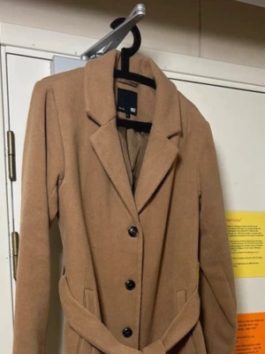 Beige kappa med bälte från lager157 - Säljer en snygg beige kappa från H&M med klassisk krage, knappar framtill och ett matchande bälte i midjan. Jackan är lång och har raka ärmar samt fickor på sidorna. Perfekt för dig som vill ha en stilren och tidlös look. Storlek L. Kamel/beige. Bra skick 