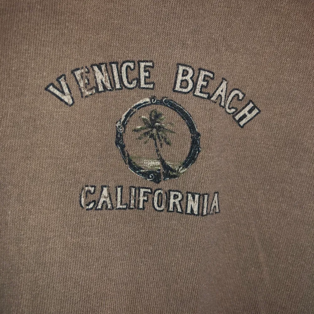 Beige/grå T-shirt med Venice Beach tryck. Litet tryck på höger framsida och stort tryck på ryggen. Uppvikta ärmar. T-shirts.