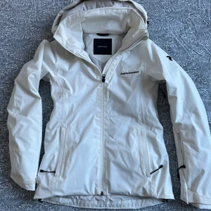 Vit Peak Performance dam W Anima J - Allround skidjacka/skaljacka. Modern slimfit-passform med smarta, praktiska fickor och blixtlåsventilation. Optimal andningsförmåga. Avtagbar luva. 