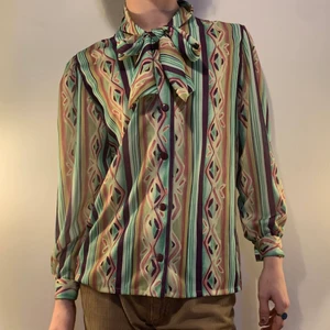 Grön lila brun gul vintage 70s skjorta - Vintage grön, lila, brun och gul 70-tals skjorta med ett psykadeliskt mönster samt två band som knyts till en rosett som lägger till en galet snygg detalj 🪲 Jag älskar också att armarna är aningen puffiga vilket gör det ännu snyggare att den är tajt vid handlederna! Jag älskar också de lila knapparna som även de lägger till en otrolig detalj! Så snygg till ett par bruna byxor för att lugna ner outfiten, ett cool disco inspirerat bälta och ett par svarta boots!