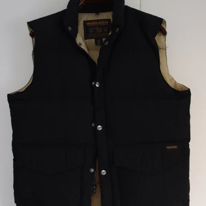 Woolrich dunväst - Svart dunväst från klassiska woolrich. Beiget foder. Är en äldre modell, men sparsamt använd. Piggen saknas från dragkedjan, men lätt att sätta dit en ny. (Se bild)