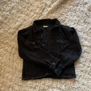 En svart zalando jeansjacka, den har två fickor där uppe och två fickor där nere, den har flera svarta knappar och den är medellång, den har även en krage där uppe, har använt hyfsat många gånger.