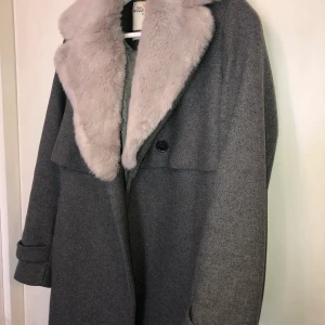 Grå kappa  - Grå kappa med avtagbar pälskrage i färgen beige/grå/rosa. Har två sidofickor, midjeskärp och knapp. Går till knäna ungefär. Knappt använd. Nypris 1299kr
