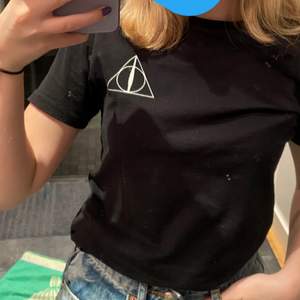 Svart t-shirt med dödsrelikerna från Harry Potter tryck på framsida och baksida. Även tillgorda hål i kanten på tröjan samt lite vid nyckelbenen. (Hålen är en del av designen) Köpt i london, fin tröja i bra skick säljer pga att den var för liten🥰 