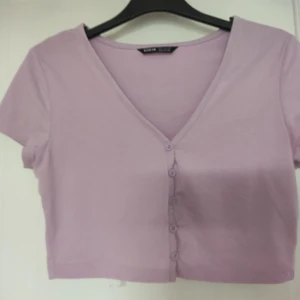 Lila kortärmad crop top med knappar - Supergullig lila kortärmad crop top med knäppning längst fram. går att ha helt uppknäppt med ett linne under, eller kombinera knäppningen fritt för många söta kombinationer.