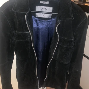 Rick Vegan Suede Black, Human Scales - Svart mocka jacka Gjord i mjukt och stretchigt Mochamaterial Den kan användas som både overshirt och som jacka   Den har även innefickor  Insidan av jackan är i en blått fint tyg 