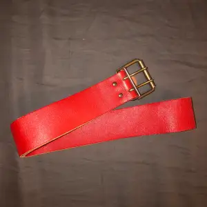 Rött läderaktigt skärp med dubbelknäppe i guldliknande metall. Längd 92cm.