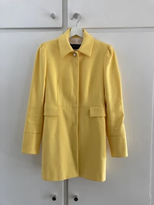 Elegant gul kappa - Superfin gul kappa med vackra knappar på ärmar och i kragen. Perfekt som en elegant kappa till finare tillfällen. Väldigt sparsamt använd och i bra skick. Säljes då den har blivit för liten för mig.