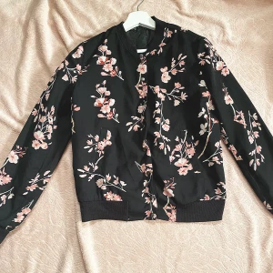 Vårjacka Blommönstrat BodyFlirt - En svart vårjacka med rosa blommönster från bodyflirt str 36. Inprincip oanvänd och inga skadar finns. Jackan har knappar som man kan stänga jackan med och extra knappar följer även med. Fickor på jackans båda sidor finns. 