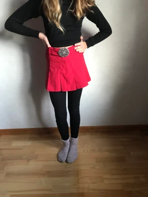 Röd snyggt kjol  - Kort kjol med snyggt metall spänne framtill. Kardborreband att ta in midjan vid behov. Storlek 146-152 som kan jämföras med en XS 